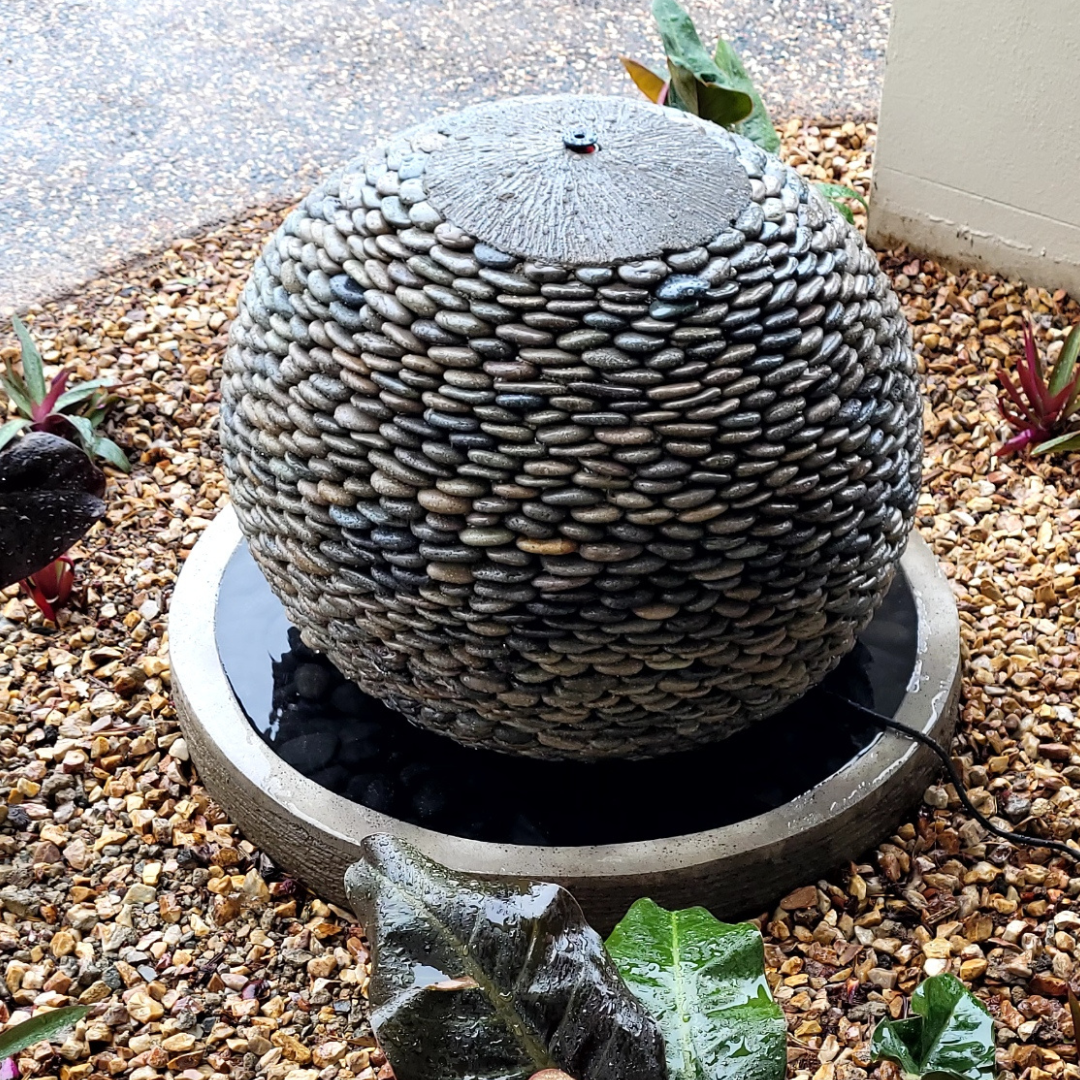 A pebble round ball water fountain with a central sphere made of pebbles and a circular base, installed in a garden setting.