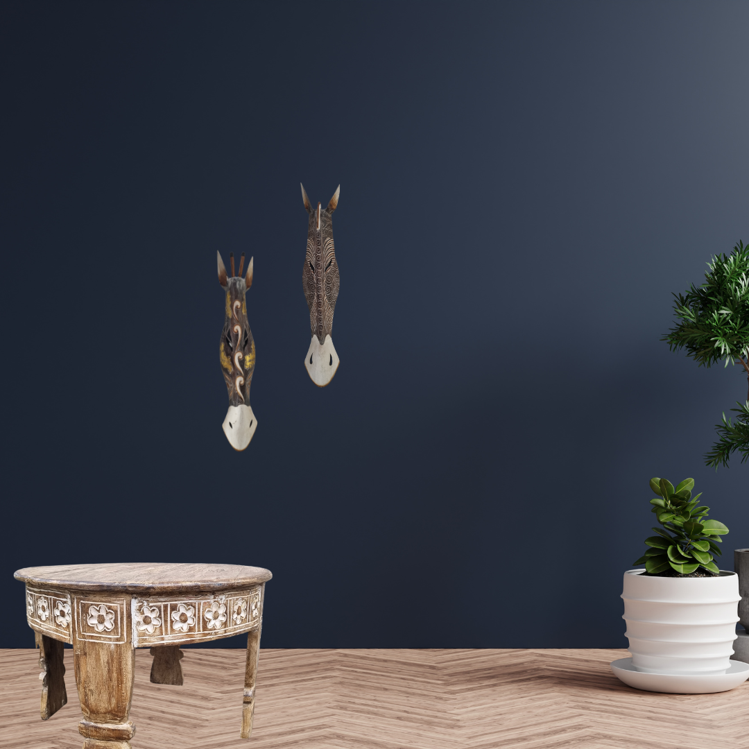 Handcrafted Wooden Giraffe & Zebra Masks Decorative wall art of animal heads on a dark blue wall with a wooden table and plant in the foreground.