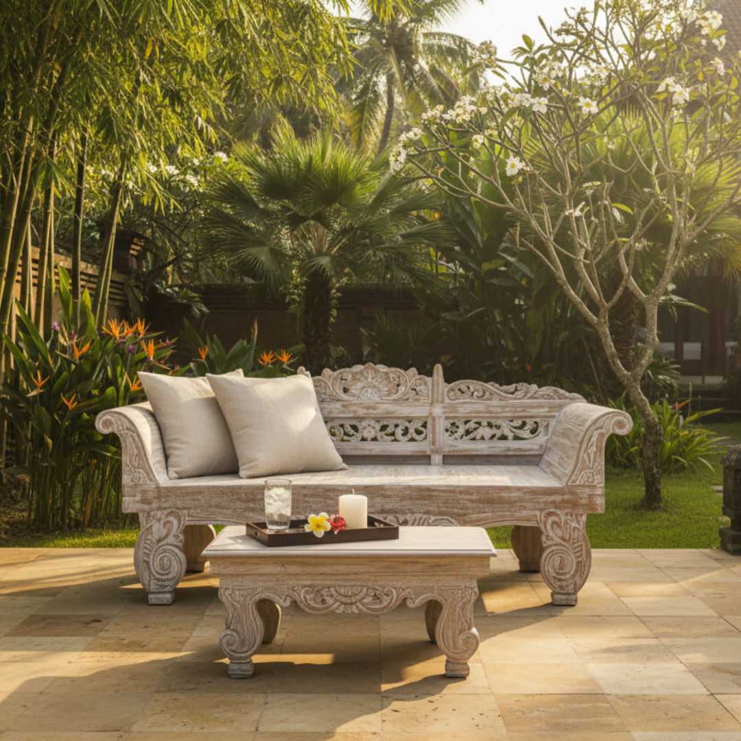 Balinese hand-carved whitewashed teak day bed with linen cushions and matching carved coffee table styled on a stone patio with tropical garden, bird of paradise flowers, frangipani tree and palm trees at golden hour – handcrafted outdoor furniture by Azure Decor