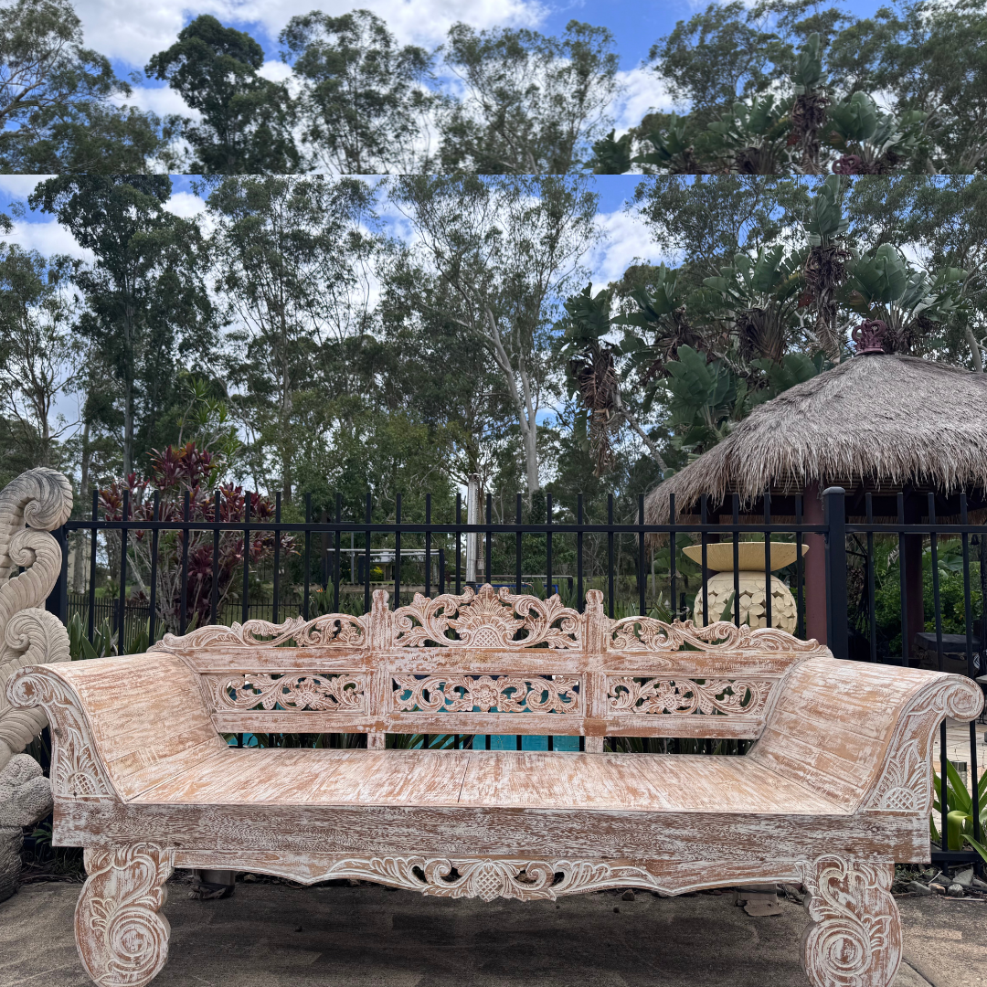 Carved wooden bench in a garden setting with trees and a thatched roof structure in the background.