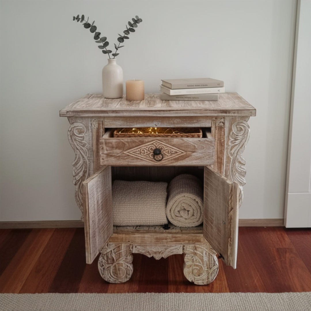 Balinese carved teak bedside table with drawer open showing fairy lights inside and cabinet doors open revealing folded blankets and a rolled towel, styled with a ceramic vase, candle, and books on top