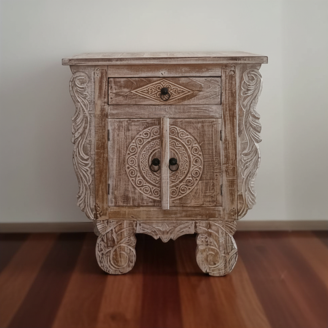Front view of a Balinese carved reclaimed teak bedside table in chalky whitewash finish on warm timber floorboards showing circular medallion cabinet doors and ornate scrollwork side panels