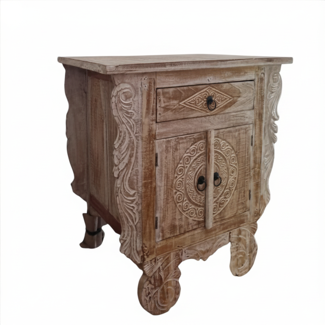 Angled product shot of a Balinese carved teak bedside table with warm sandy whitewash finish showing scrollwork side panels, circular medallion double doors, diamond drawer, and decorative bun feet