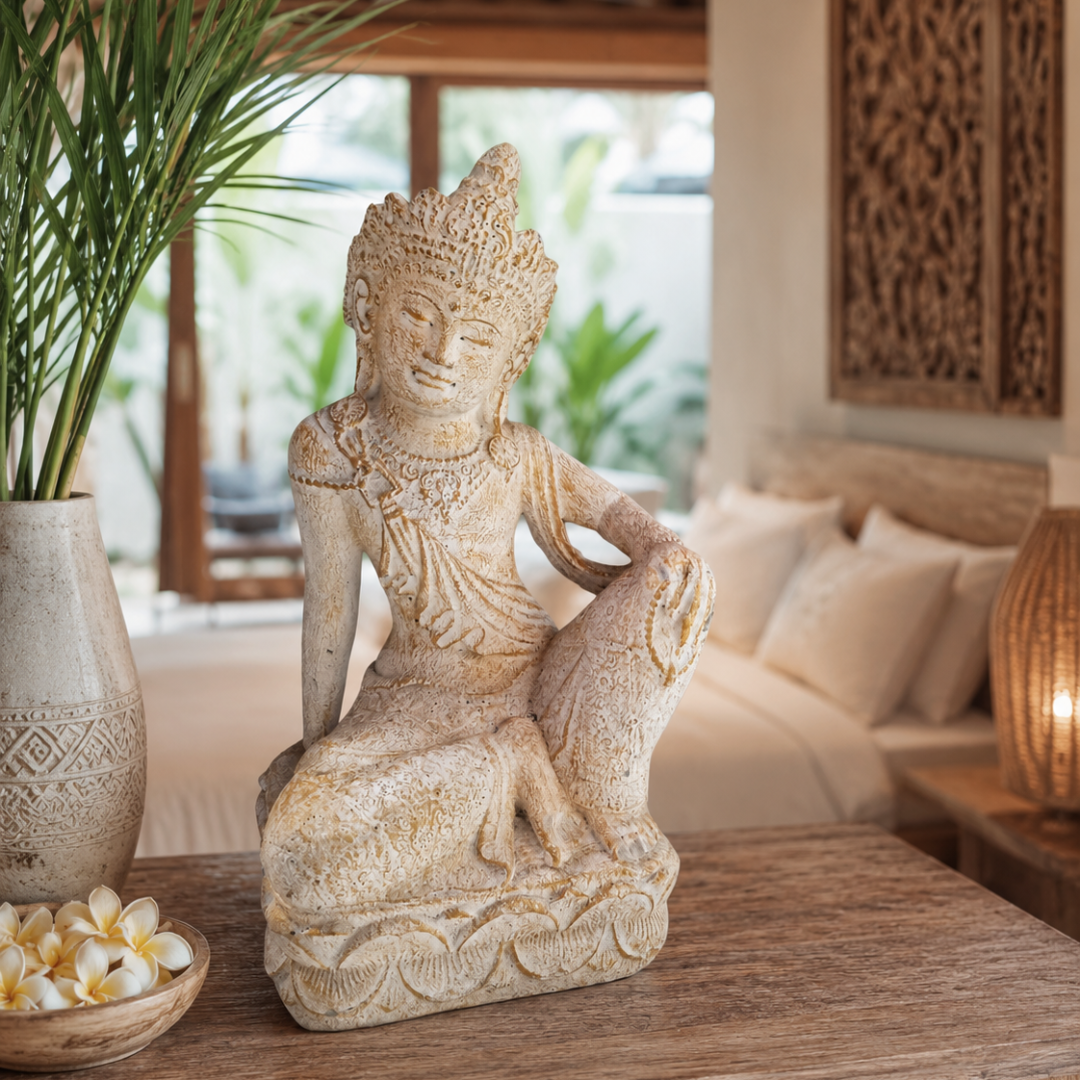 Seated Balinese goddess statue with relaxed open pose and carved robes styled on a carved timber coffee table beside a white ceramic vase with palm leaves and a bowl of frangipani flowers