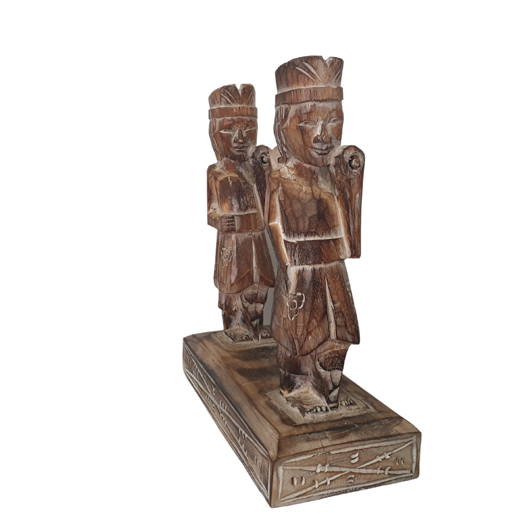A wooden statue depicting two Balinese figures in traditional ceremony attire, standing on a platform.