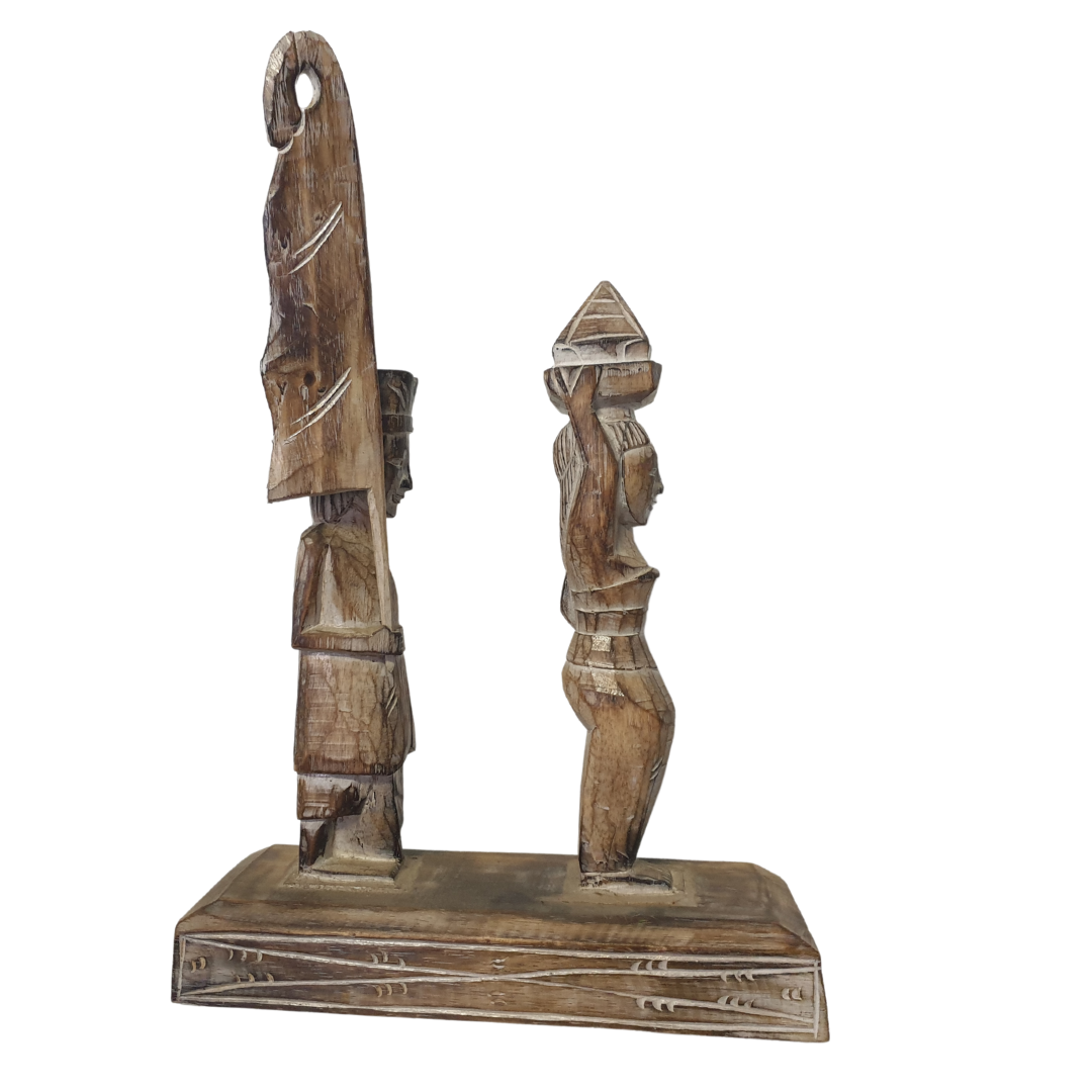 A wooden statue depicting two Balinese figures in traditional ceremony attire, standing on a platform.