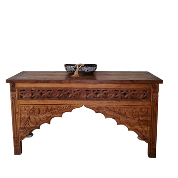 A Balinese style console table made of rustic teak wood, featuring intricate carvings and a bowl on top, with a dark brown finish.