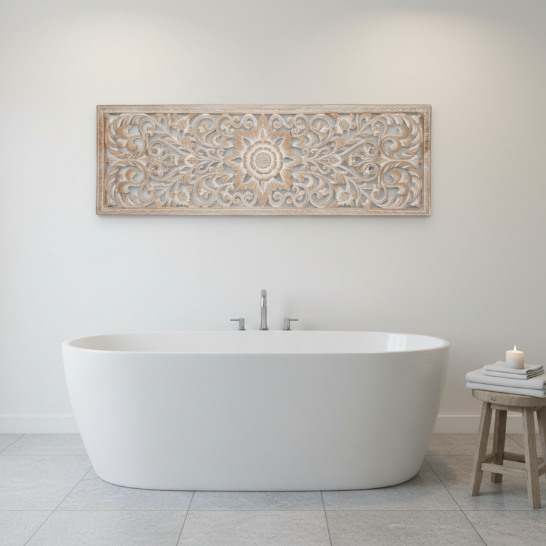 Whitewash precision-cut MDF wall art panel with Balinese floral scroll design hung horizontally above a freestanding oval bathtub in a bright all-white luxury bathroom, with a timber stool, candle and stacked books beside the bath
