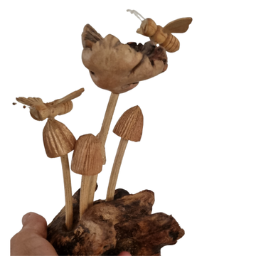 A hand-carved wooden fairy garden figurine featuring mushrooms and a bee on a driftwood base, held by a hand against a white background.