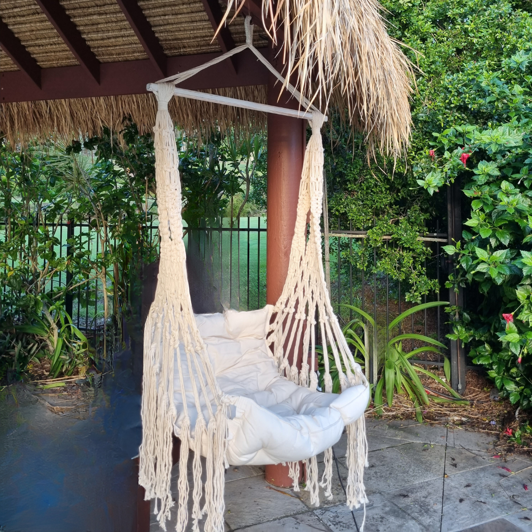 A beige cotton Boho Macrame hammock chair hanging under a thatched roof with surrounding greenery.