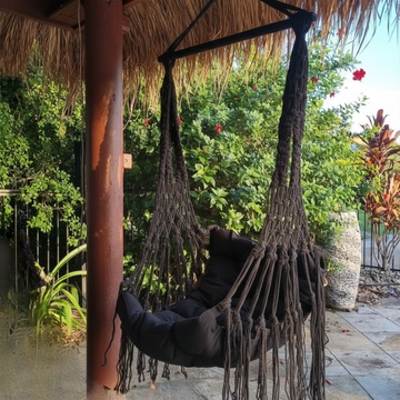 Black hammock swing hanging from a thatched roof with greenery in the background