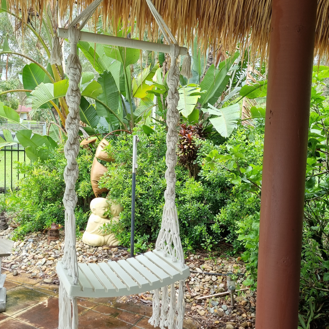A handcrafted Balinese style white macrame swing with a solid wooden seat, designed with intricate patterns and tassels.