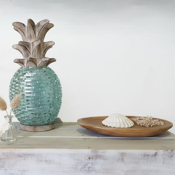 Boho Glass Pineapple Décor in aqua sea-glass with whitewashed carved wooden leaf top, styled as the hero piece on a whitewashed timber shelf with warm side lighting catching the glass texture, a small rustic wooden tray, dried botanicals in a bud vase and a single white shell — coastal boho tabletop sculpture by Azure Decor