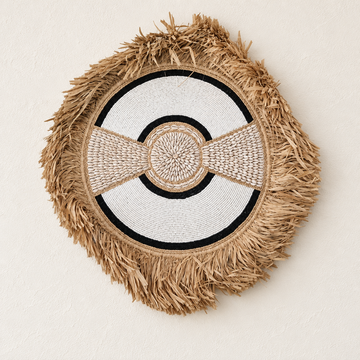 Black-accent circular wall hanging with shell centre and straw fringe on a warm white wall