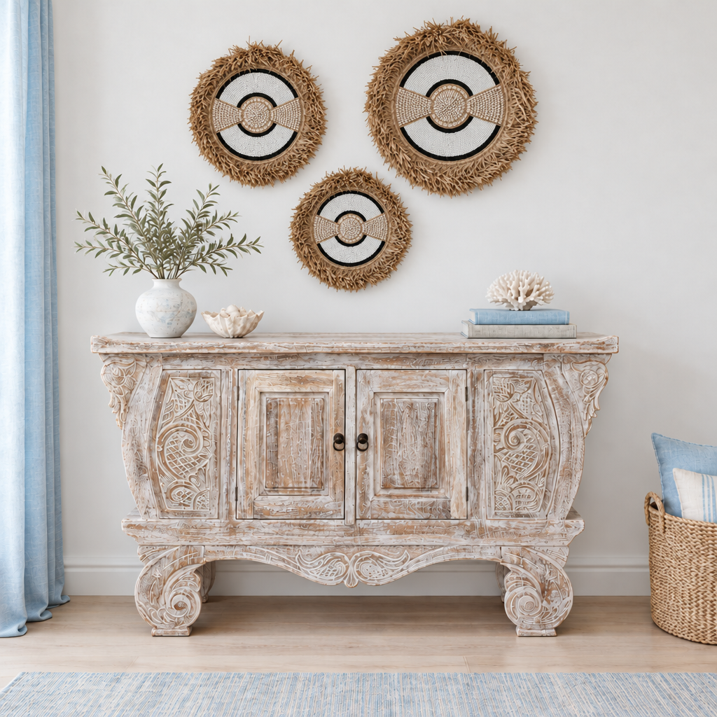 Three black-accent shell-and-bead shields arranged above a weathered carved cabinet in a coastal room.
