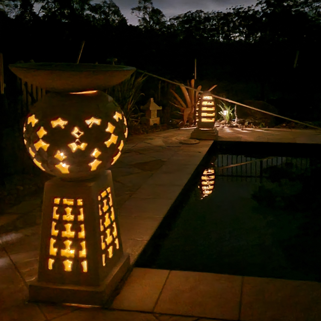 Bola Style Garden Birdbath lit from within at night beside a pool, casting warm golden light through the carved frangipani sphere