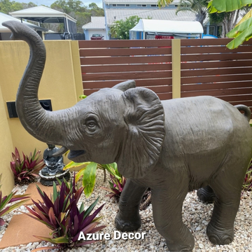 Brown elephant statue water feature with a raised trunk, standing on white gravel among tropical garden plants.