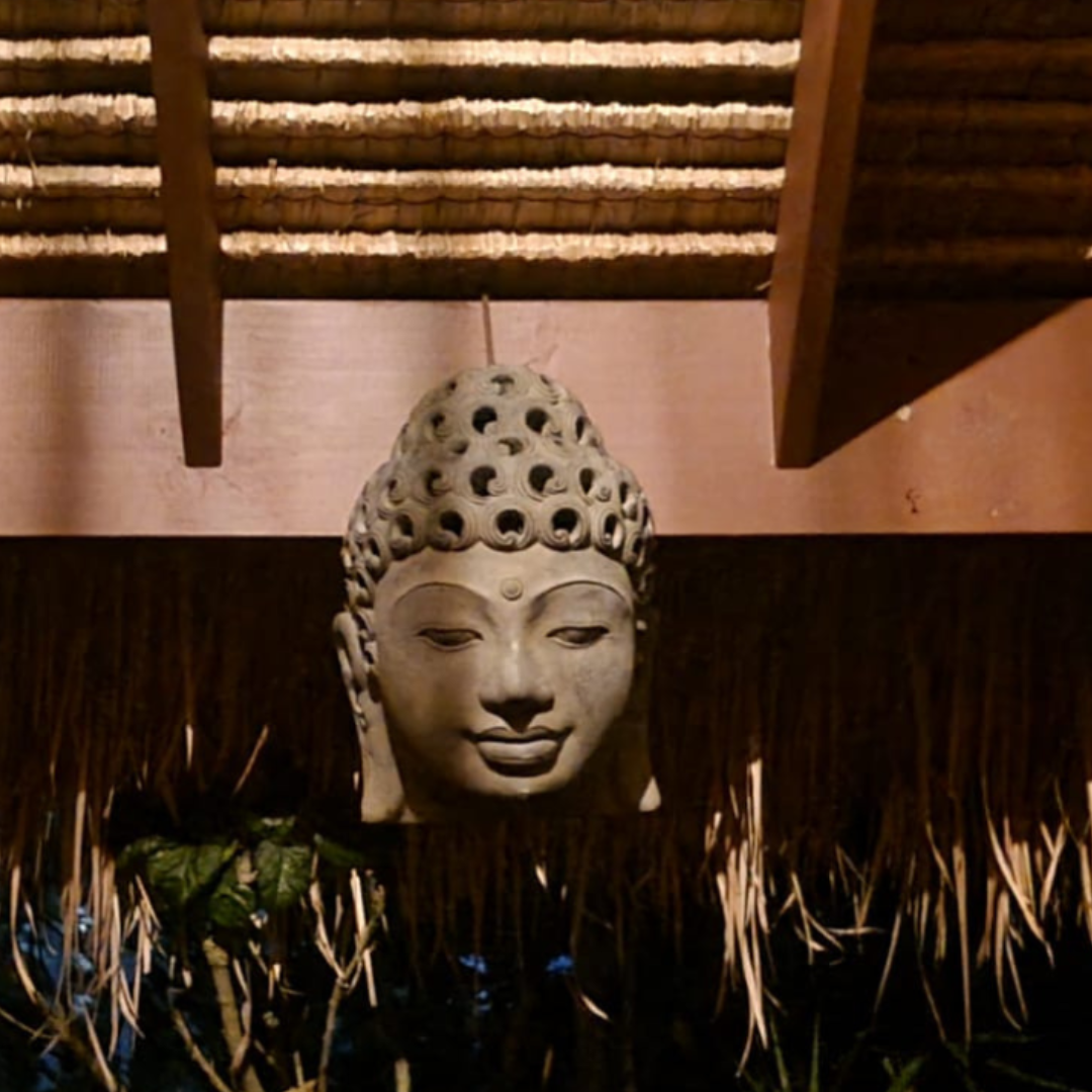 Grey Buddha head wall plaque displayed on a dark outdoor wall beneath a thatched roof, surrounded by tropical plant