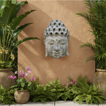 Silver-grey Buddha head wall plaque mounted on a textured outdoor wall surrounded by tropical plants and potted flowers.