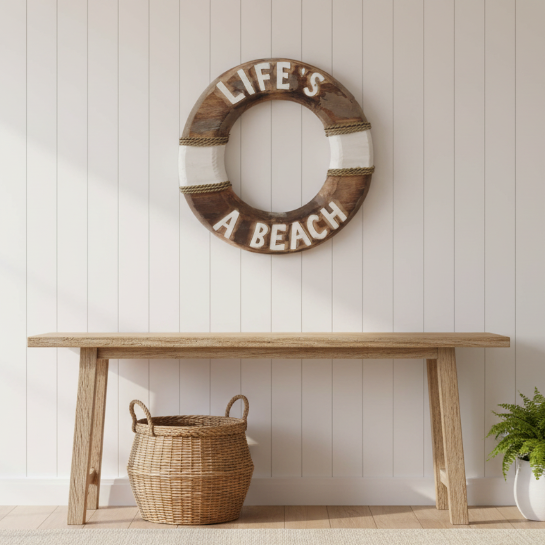 A rustic wooden wall sign reading 'LIFE'S A BEACH' in raised white letters on a distressed brown and white life preserver shape, mounted on a white paneled wall above a light wood bench with a woven basket underneath.