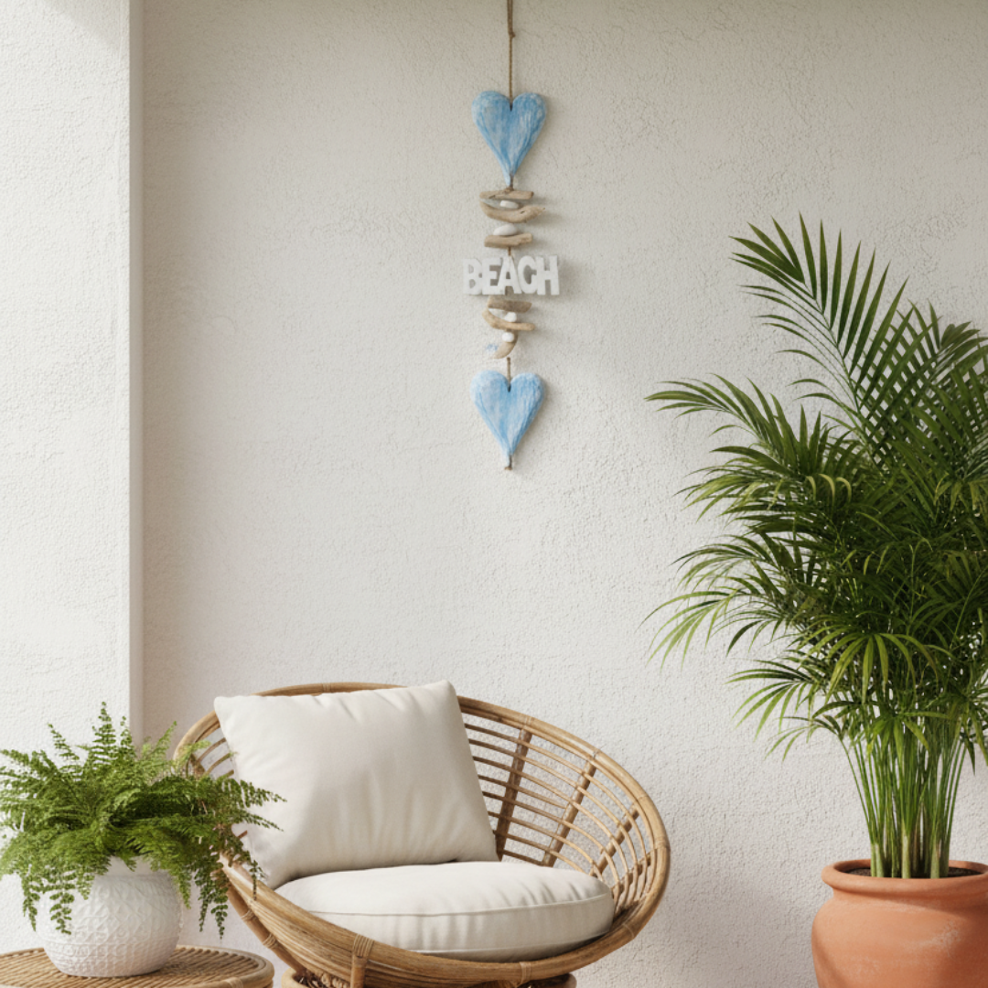 A decorative hanging wall ornament with two light blue heart shapes, small shells, and a white sign reading “BEACH,” displayed on a textured white wall above a wicker chair with white cushions, alongside potted green plants.