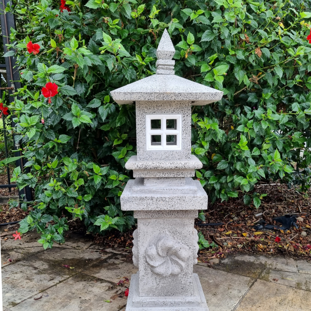 Tall light grey outdoor sculpture with carved spiral detail on the column, square window opening and pointed finial, styled against a lush green hibiscus hedge