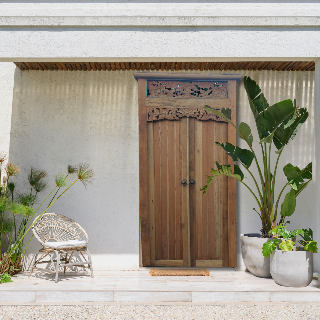 Wooden door with decorative elements on a light gray wall, surrounded by plants and a chair.