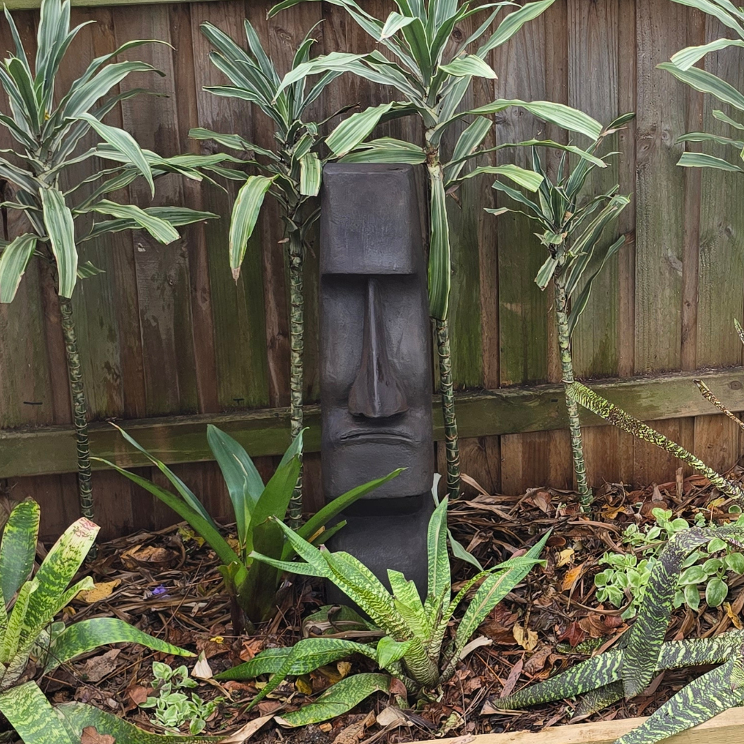 HEAD STATUE EASTER ISLAND STYLE in a garden setting with plants and a wooden fence.