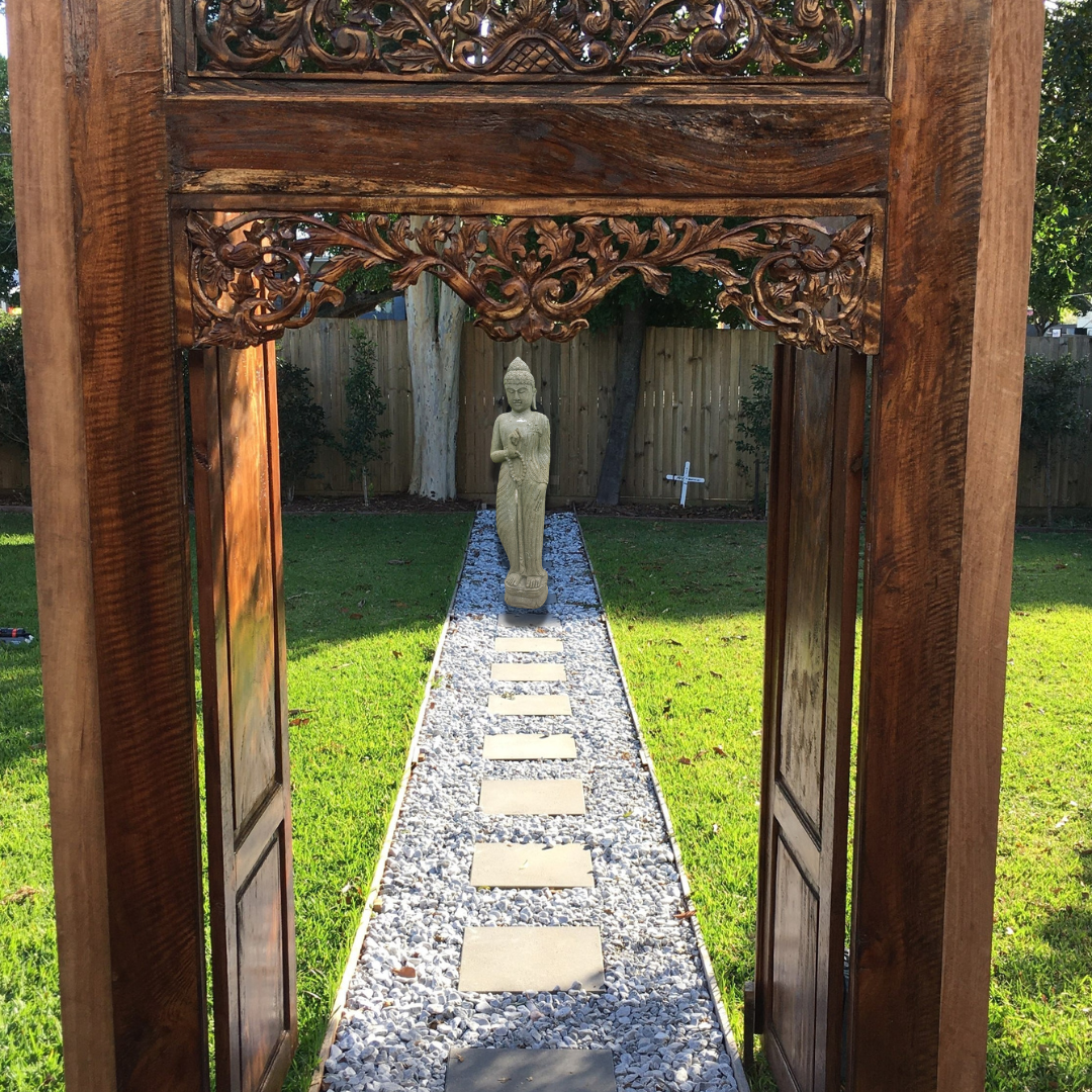A carved wooden garden archway framing a stone path with stepping stones leading to a in a grassy backyard.