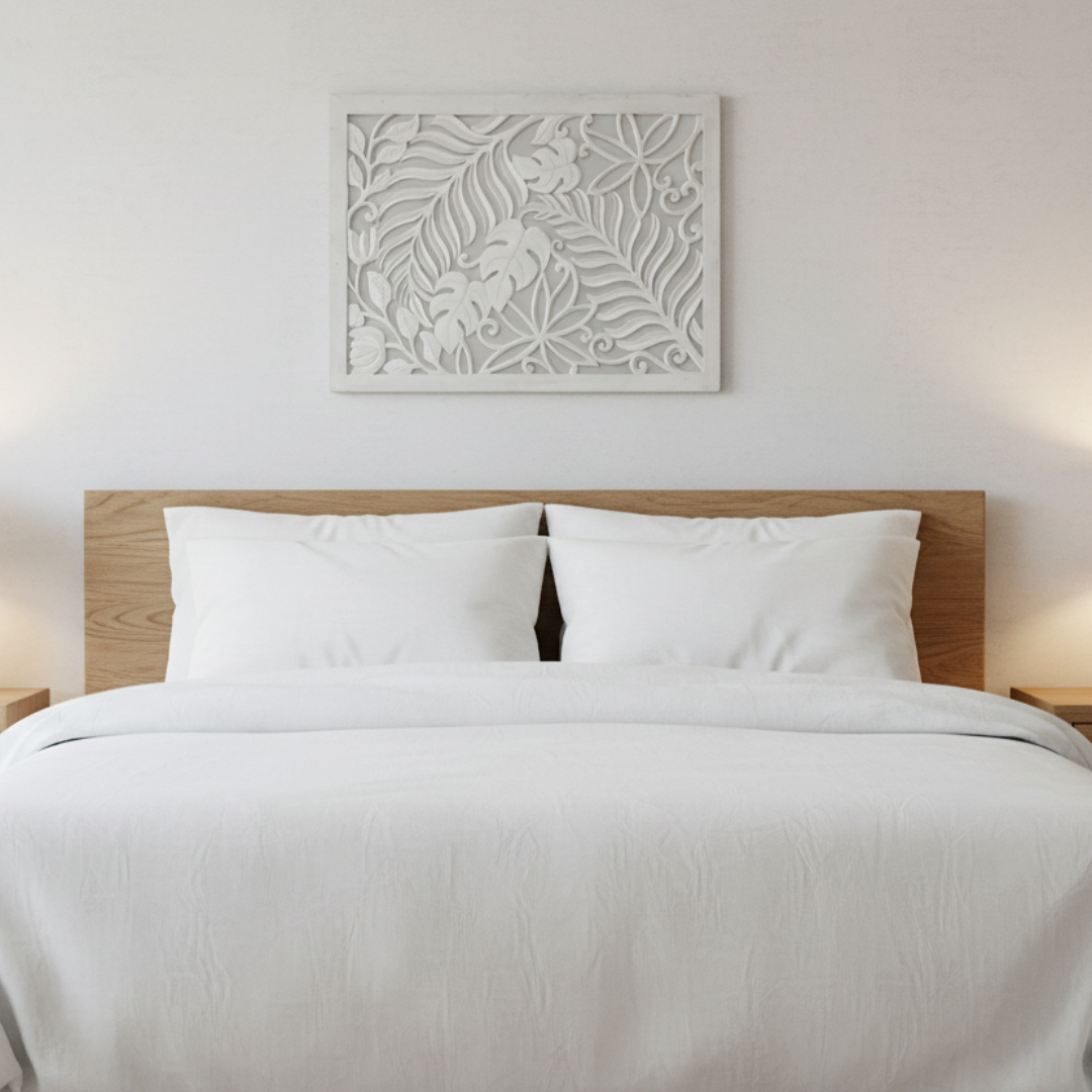 Whitewash carved timber wall art panel hung horizontally above a timber bed headboard in a serene white bedroom, flanked by warm bedside lamps and crisp white linen bedding