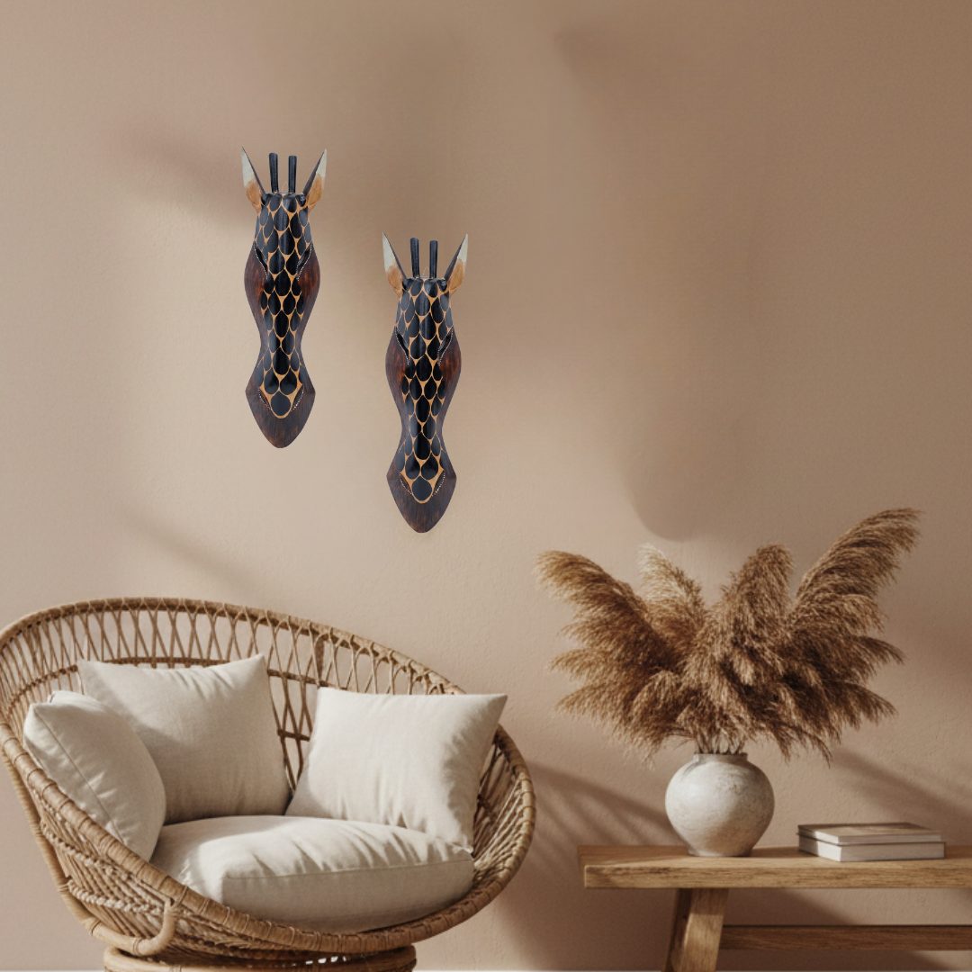 Two dark blue and brown giraffe wall art pieces with gold accents hang on a beige wall above a wicker chair and a side table with dried grass in a vase.