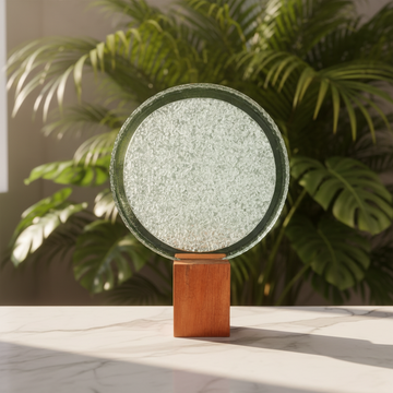 Circular clear disc on timber base on a white marble surface with soft natural light and lush tropical palm foliage
