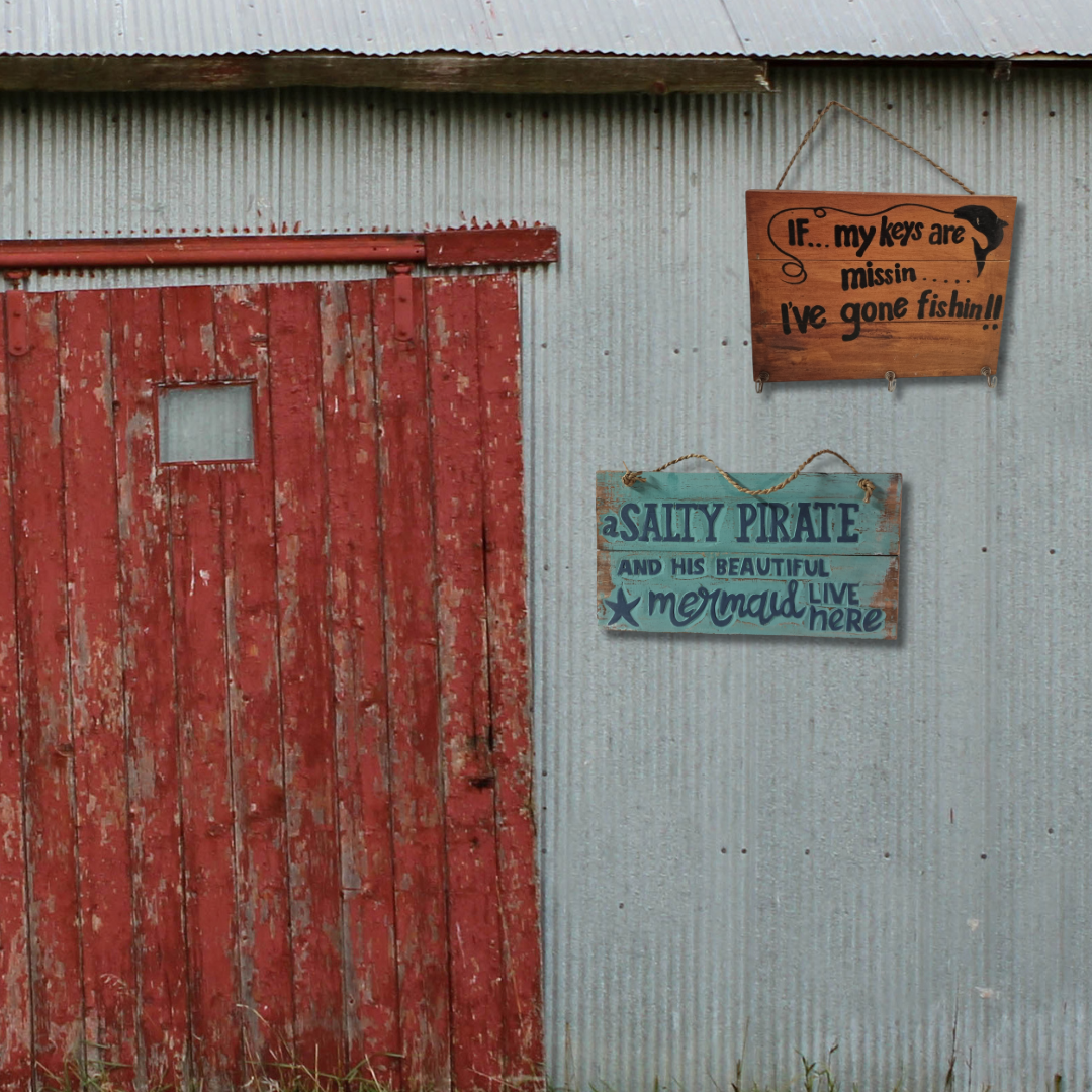 Gone Fishing and A Salty Pirate wooden wall plaques hung side by side on a rustic corrugated iron shed wall beside a weathered red barn door