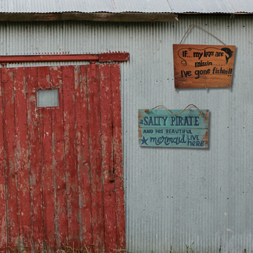 Gone Fishing and A Salty Pirate wooden wall plaques hung side by side on a rustic corrugated iron shed wall beside a weathered red barn door