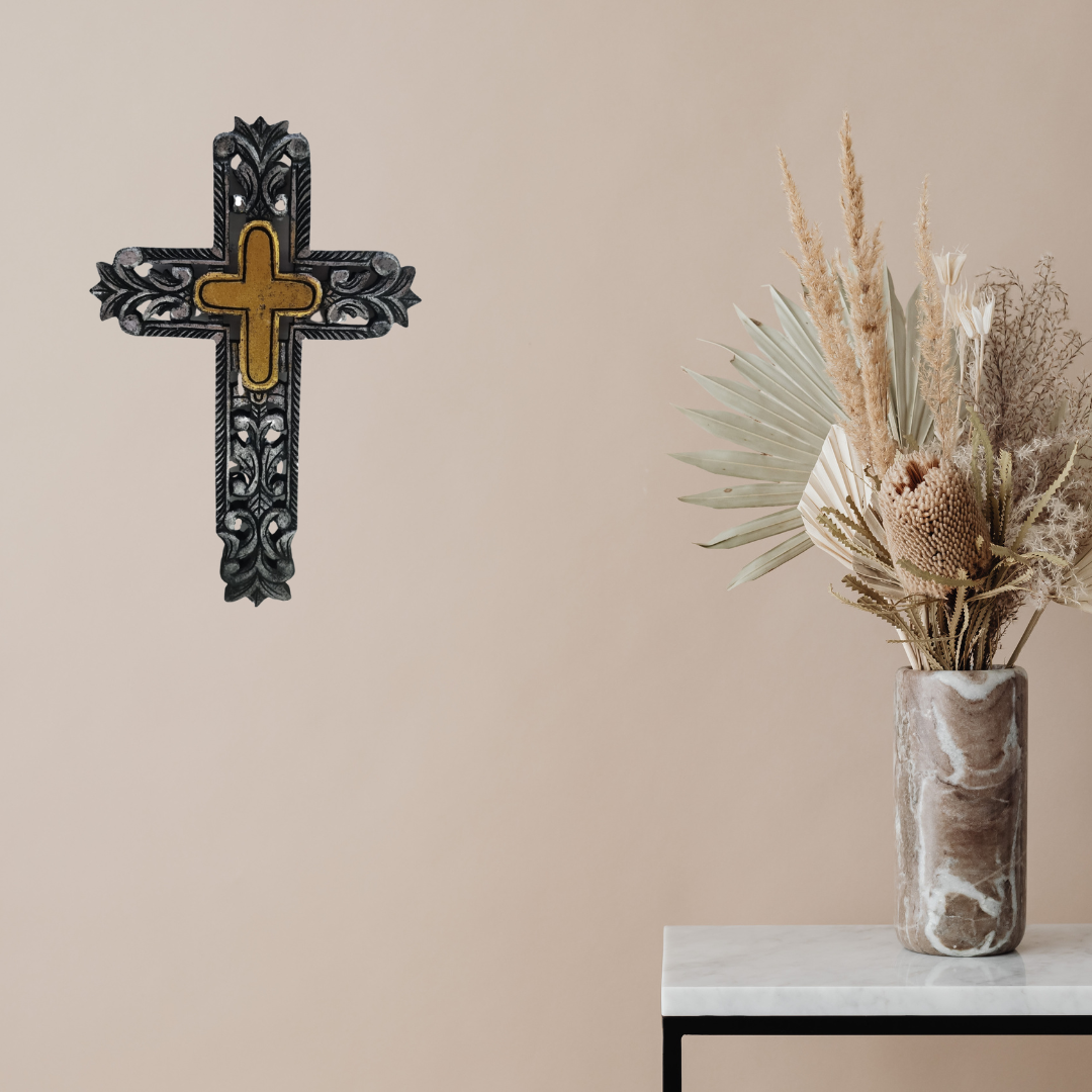 Decorative cross on a wall next to a vase with dried plants on a table.