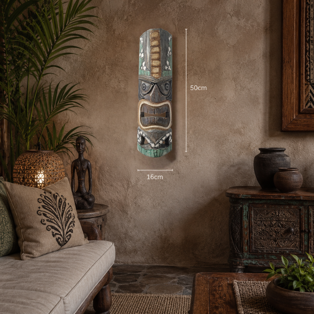 Hand-carved wooden tiki mask with angular headdress and gold detailing hung on a warm textured plaster wall in a tropical living room, styled with a cream linen sofa, rattan pendant lamp, palm plant, and carved timber cabinet.