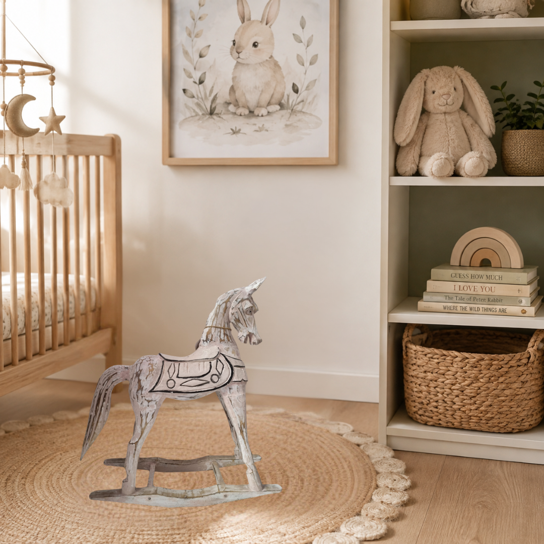 A styled nursery corner featuring a handcrafted rustic rocking horse white-painted wooden on a round woven jute rug, next to a light wood crib and a shelving unit with a plush bunny toy and framed rabbit artwork.