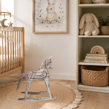 A styled nursery corner featuring a handcrafted rustic rocking horse white-painted wooden on a round woven jute rug, next to a light wood crib and a shelving unit with a plush bunny toy and framed rabbit artwork.
