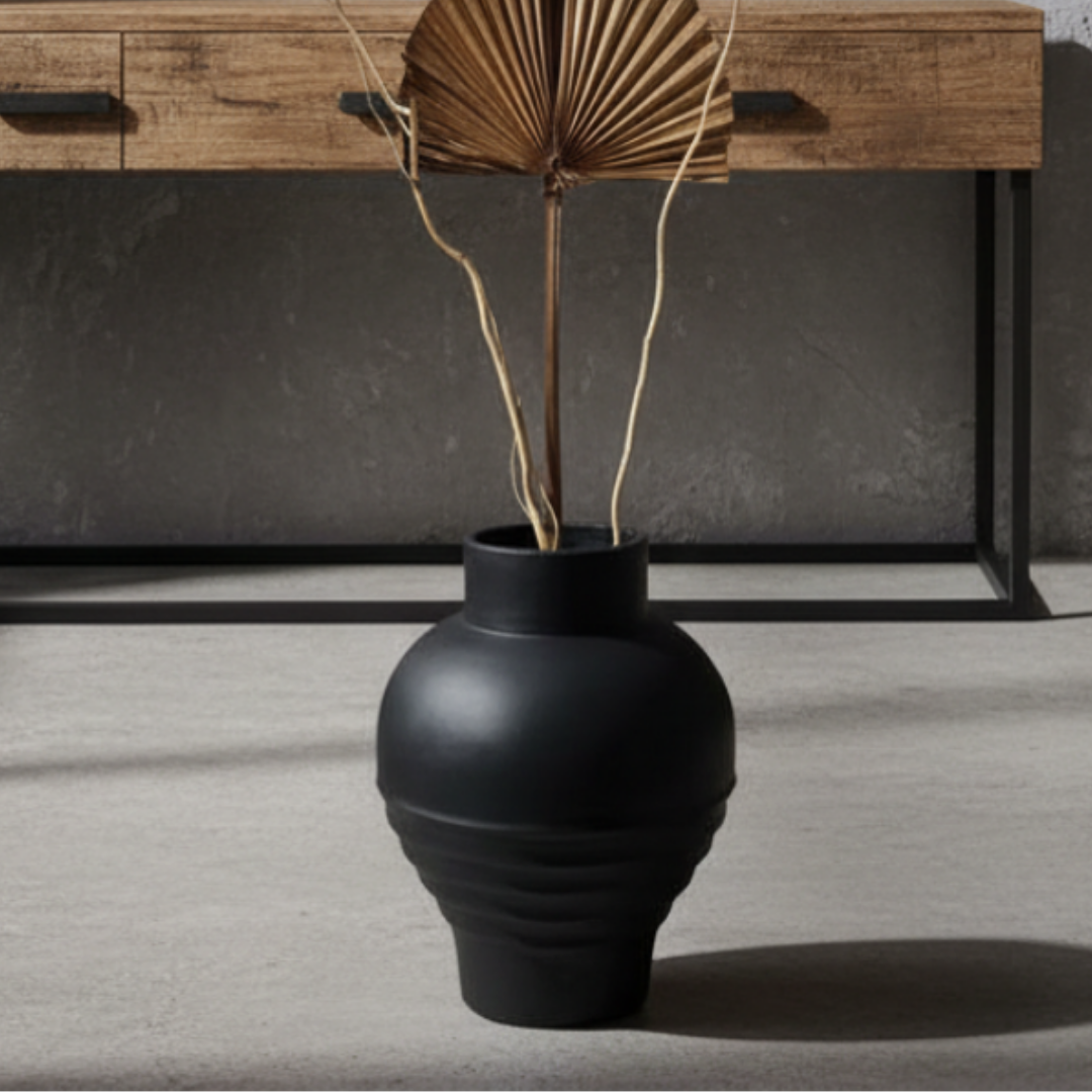 A matte black vase with a ribbed textured base, holding a dried palm leaf stem, set on a light gray floor in front of a dark gray wall and a wooden console table.