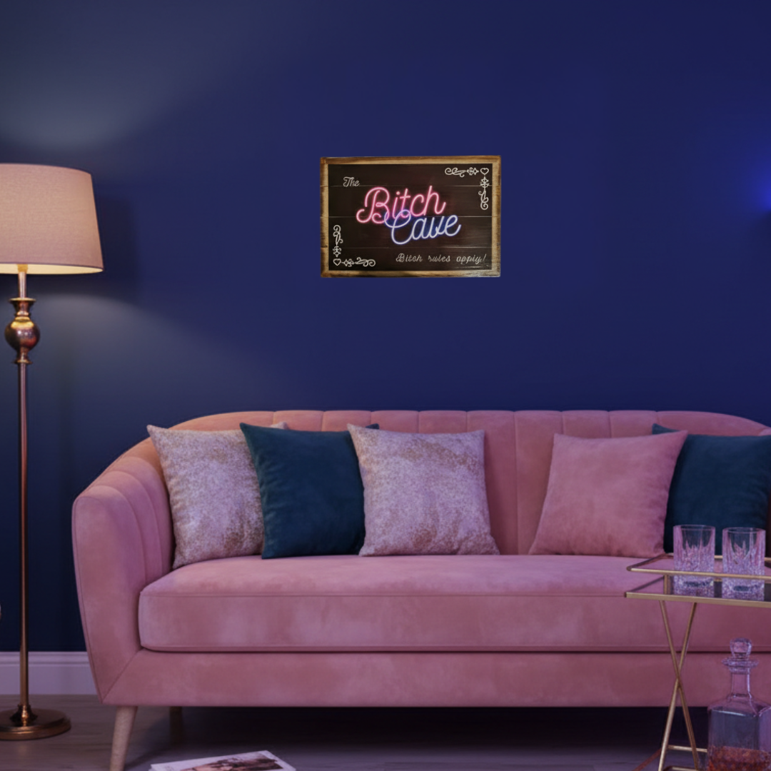 A framed wall sign reading 'Bitch Cave' in pink and white script on a dark background, mounted above a pink sofa with blue and beige pillows in a styled room with a blue wall, floor lamp, and side table.