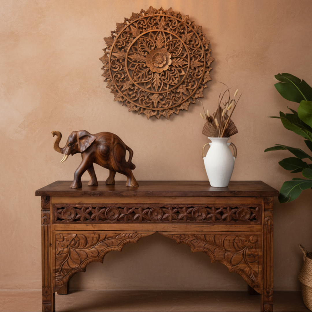 Dark warm brown hand-carved figurine with trunk raised styled on an ornate carved timber console table, beside a white ceramic vase with dried palm leaves and botanicals, with a large round carved wooden mandala panel on the wall above, in a warm Balinese-modern hallway setting