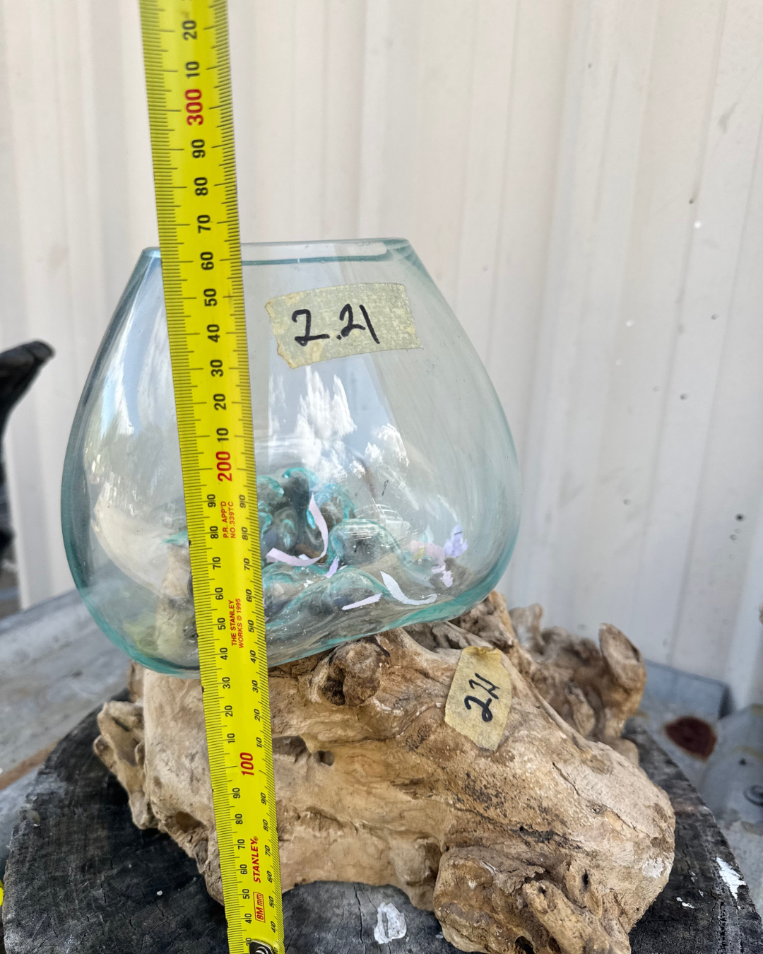 A small teardrop-shaped clear glass vase or planter displayed on a piece of, with a yellow tape measure standing alongside it and handwritten price tags reading 2.4 visible