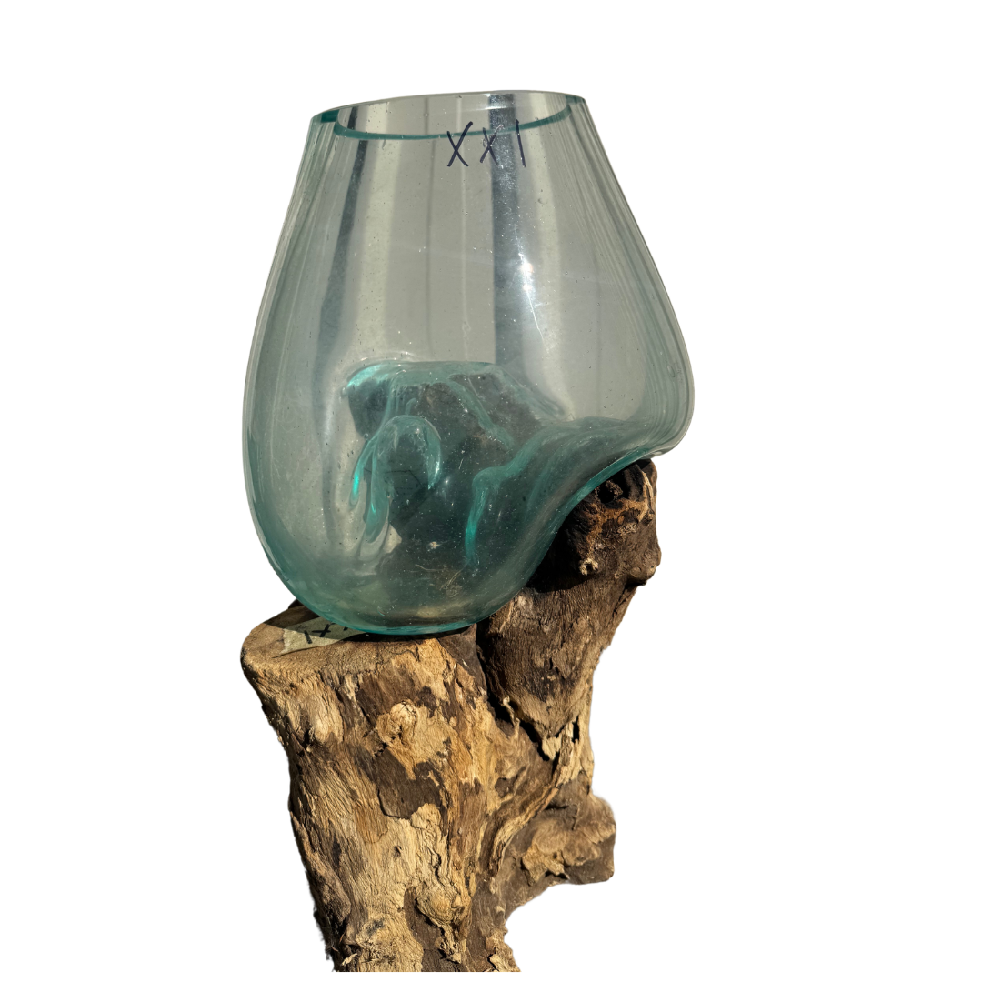 A translucent smoky blue-green glass bowl or vase with a sculptural organic shape, displayed on a natural driftwood stand against a plain white background.