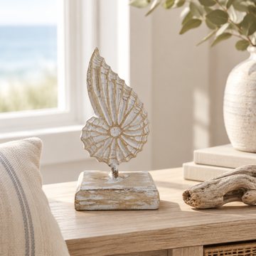 A carved shell-shaped ornament on a timber base, styled on a console shelf beside a linen cushion and a piece of driftwood with an ocean view through the window.