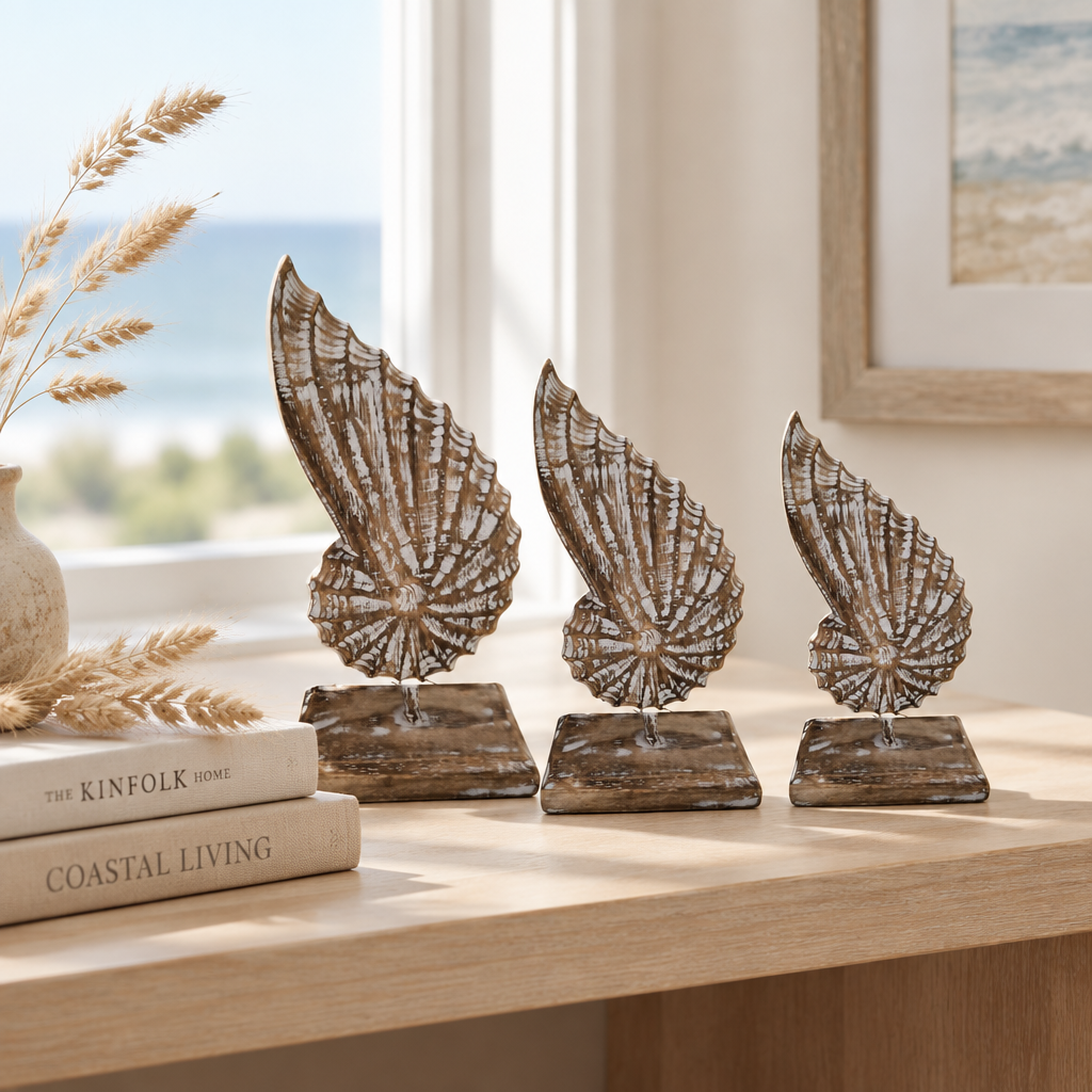 Three carved shell-shaped ornaments in graduated sizes, styled on a timber console shelf with a stack of books and dried coastal grass, ocean view through the window.