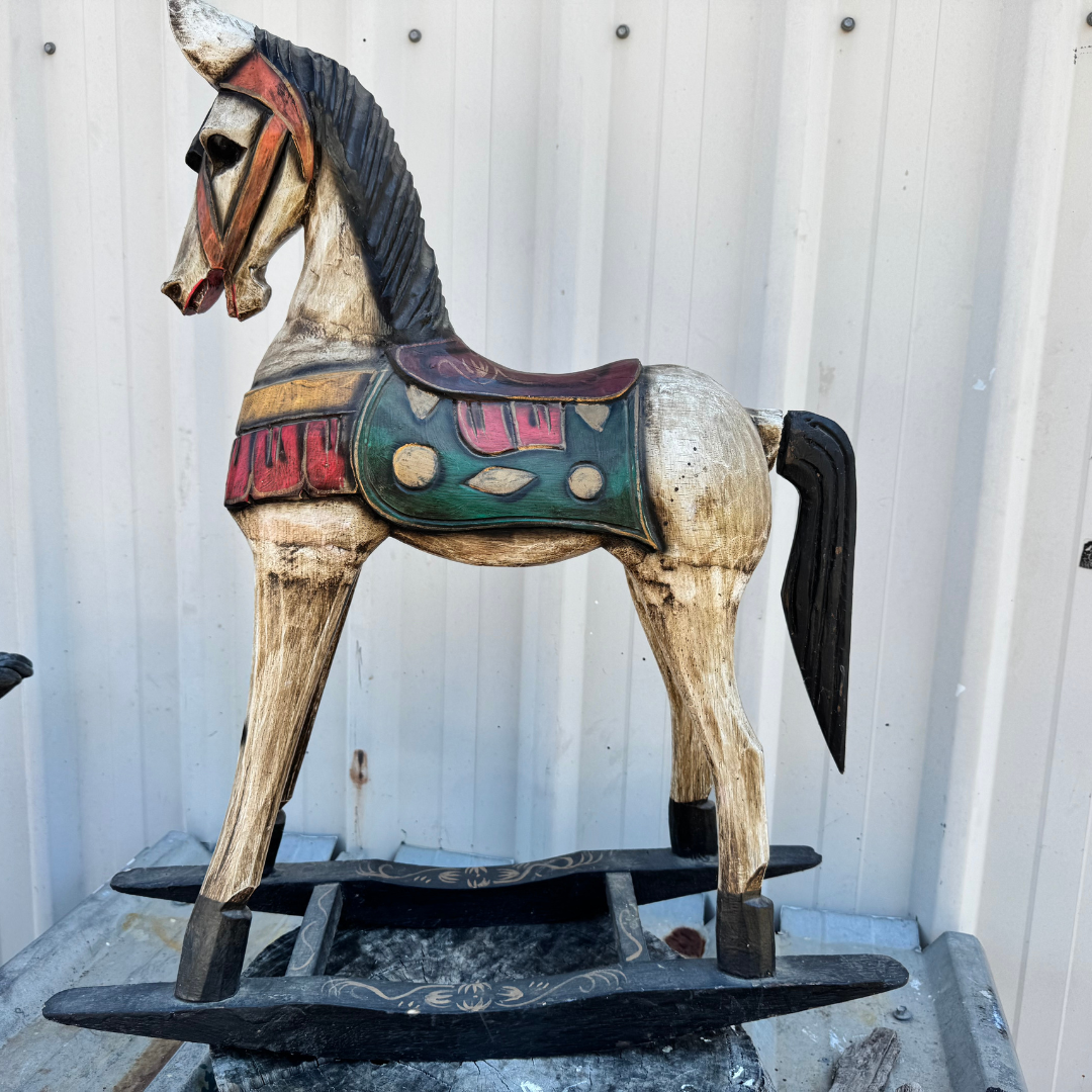 Rustic timber horse sculpture with weathered tan body, lighter blue and green saddle detailing, gold accents, and black rocker base, against a corrugated iron wall.