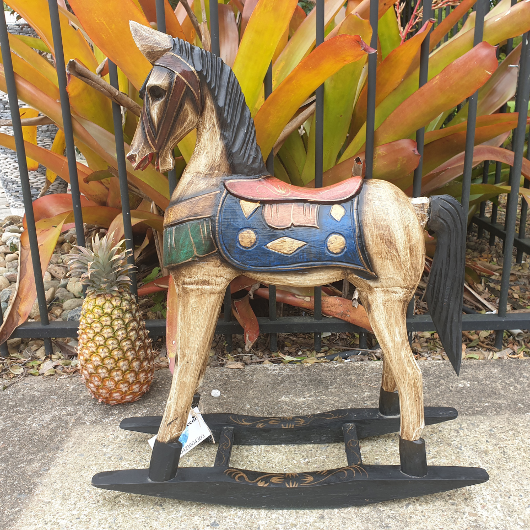 Hand-carved timber horse with weathered tan body, bold blue and red saddle, green trim, and black rocker base, photographed outdoors against tropical bromeliad plants and iron fencing