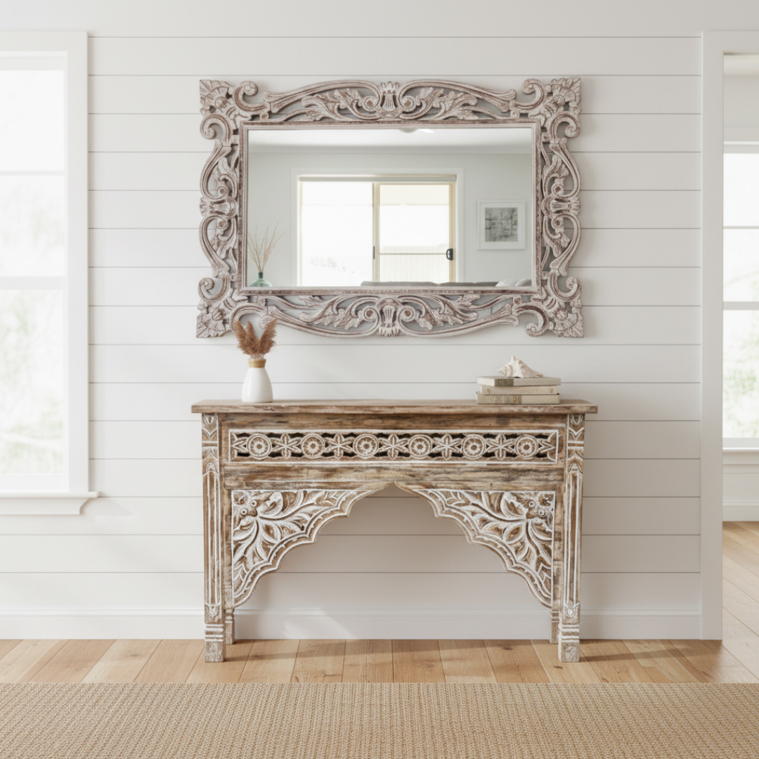 Decorative wooden console table with ornate carvings and a matching mirror in a room with white walls and wooden flooring.