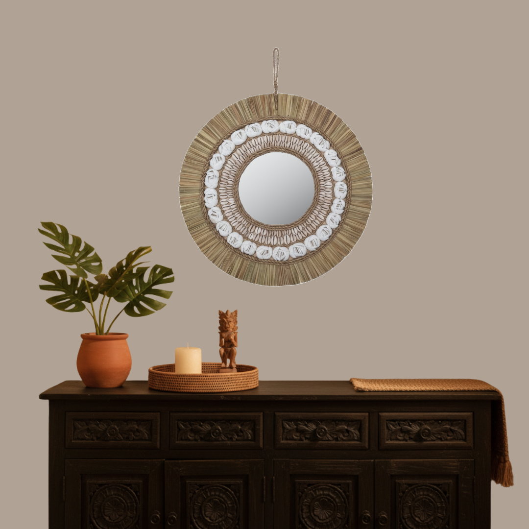 Natural tone boho coastal wall mirror with raffia fringe and cowrie shell detail, styled above a dark carved Balinese timber cabinet with monstera plant, candle and wooden figurine — 40x40cm — handcrafted coastal boho home decor by Azure Decor