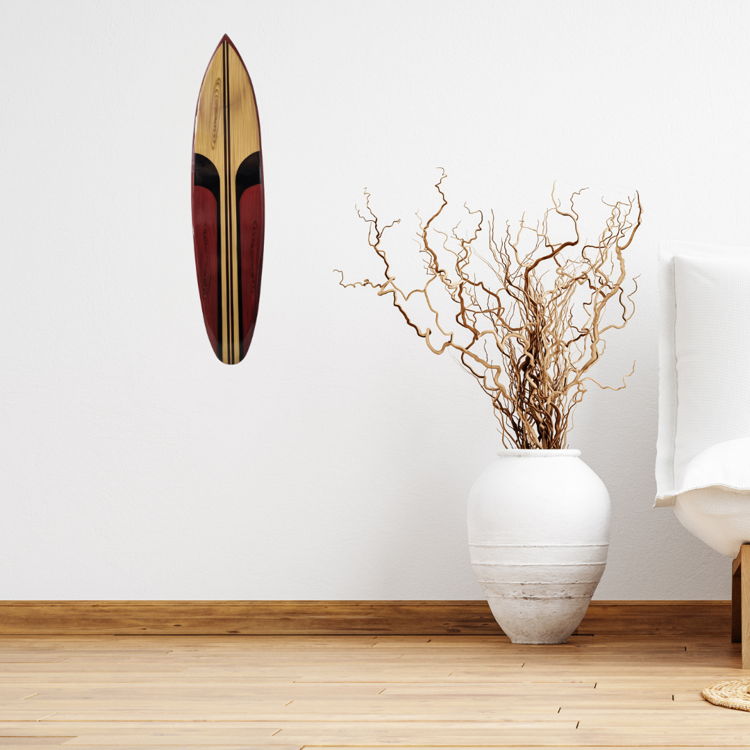 Wooden surfboard wall art above a white vase with branches on a wooden floor.